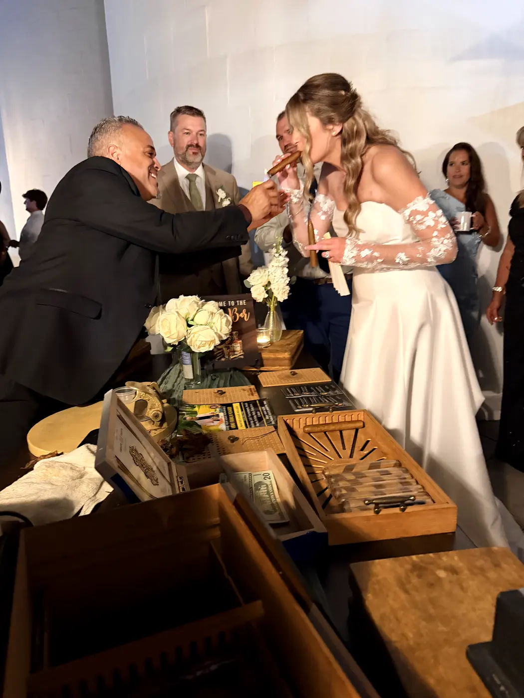 The bride at his table taking a lit cigar, the groom beside her and guests gathered behind