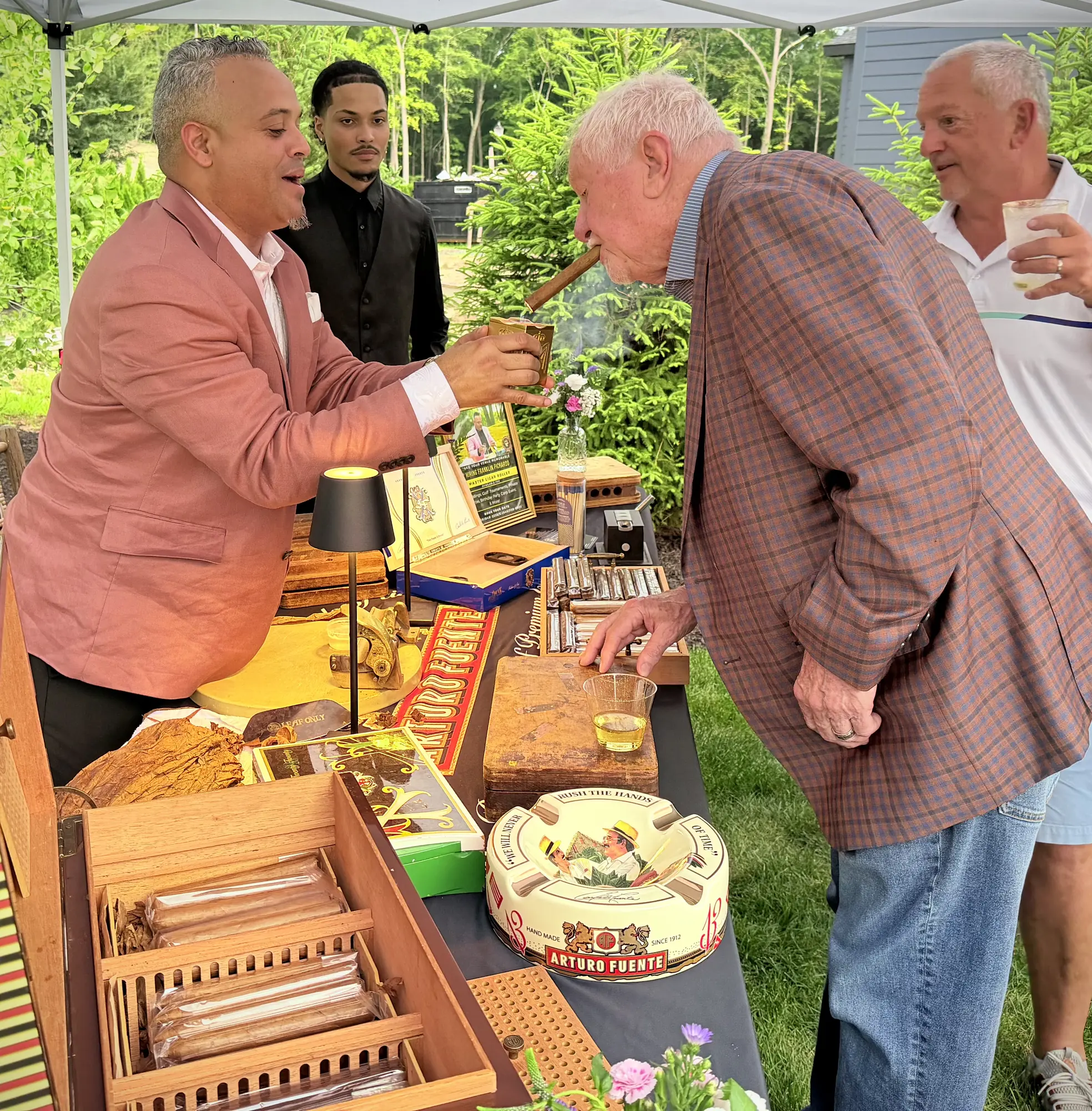 Franklin lighting a cigar for a guest at his table on a lawn, with other guests around him